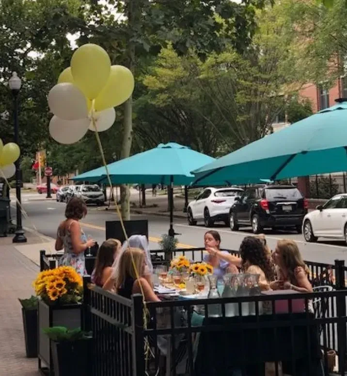 Group of women dining outdoors with sunflowers and teal umbrellas.