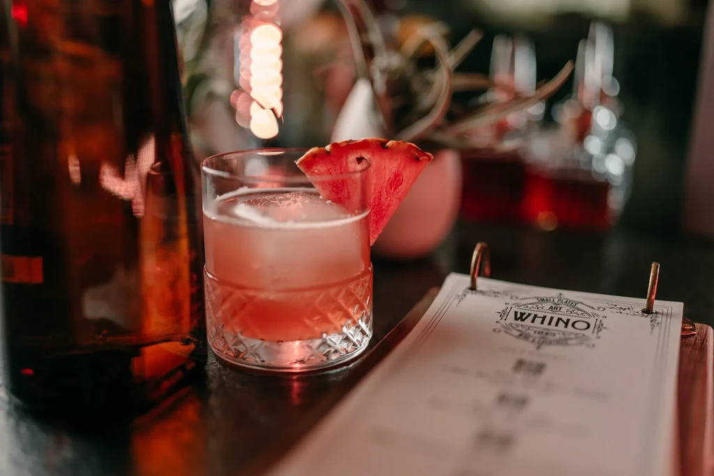 Close-up of pink cocktail, watermelon, and "WHINO" menu card.