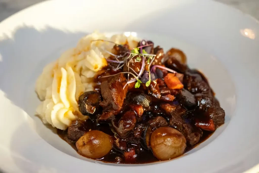 Plated meat stew with mashed potatoes, pearl onions, mushrooms, and microgreens.
