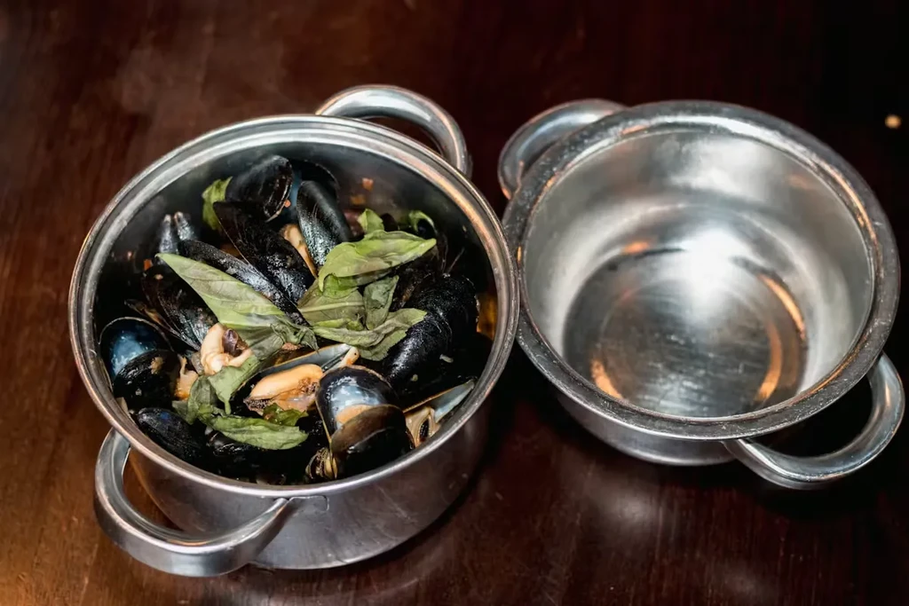 Two silver pots, one with steaming mussels and basil, one empty.