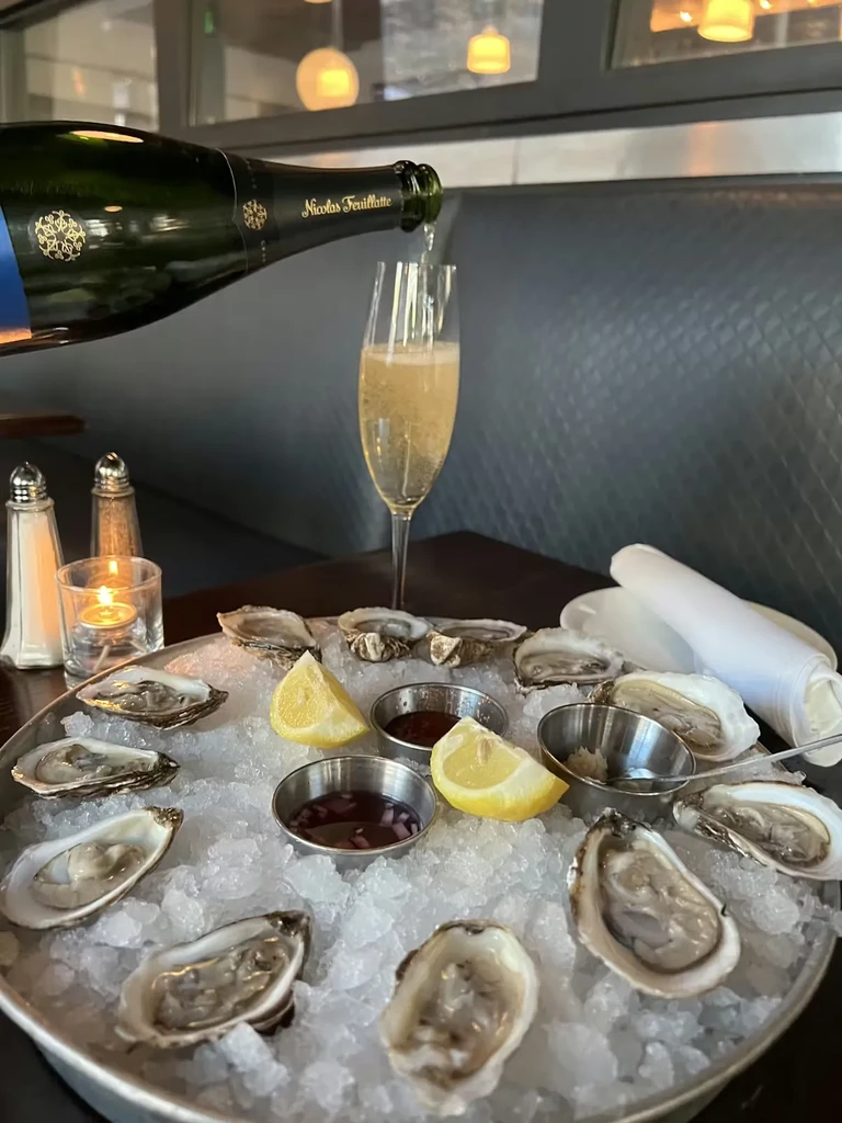 Raw oysters on ice with sauces, lemon, and champagne pouring.