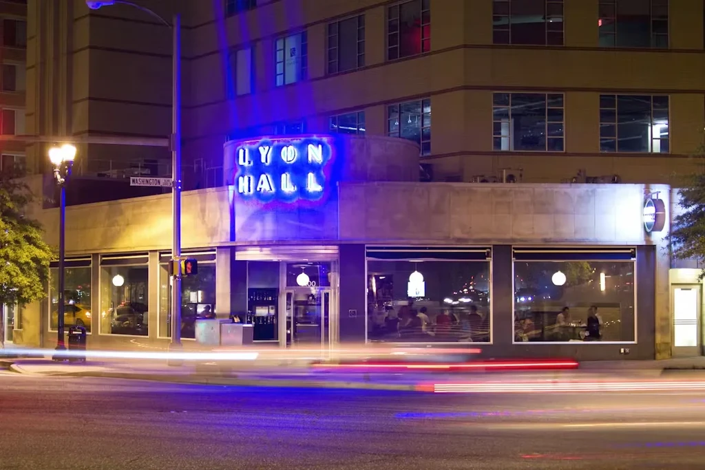 Nighttime view of Lyon Hall restaurant with blue neon sign and vehicle light trails.