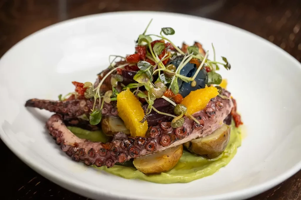 Charred octopus tentacle on potatoes with green purée and colorful garnishes.