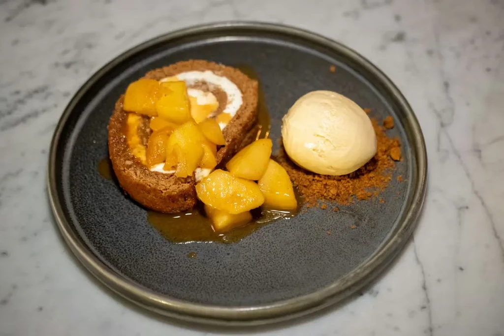 Cake roll with fruit, sauce, and ice cream on dark plate.