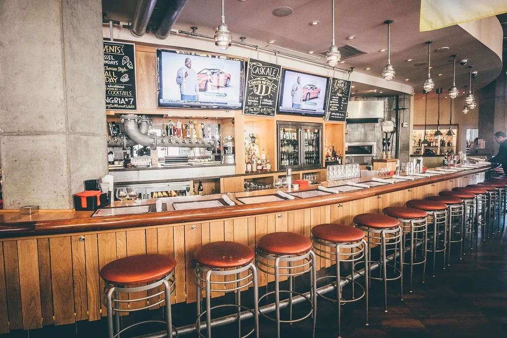 Modern industrial bar with curved wooden counter, red stools, and TVs.