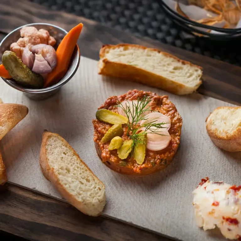 Appetizer platter with steak tartare, bread, pickled vegetables, and creamy spread.