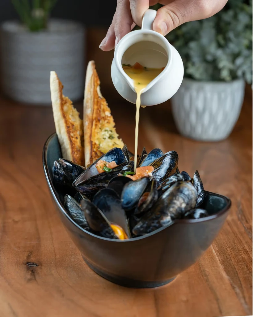 Hand pouring creamy sauce over mussels and bread in a bowl.
