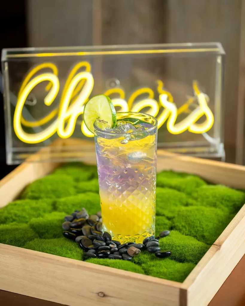 Colorful layered drink with lime on moss, with a "Cheers" neon sign.