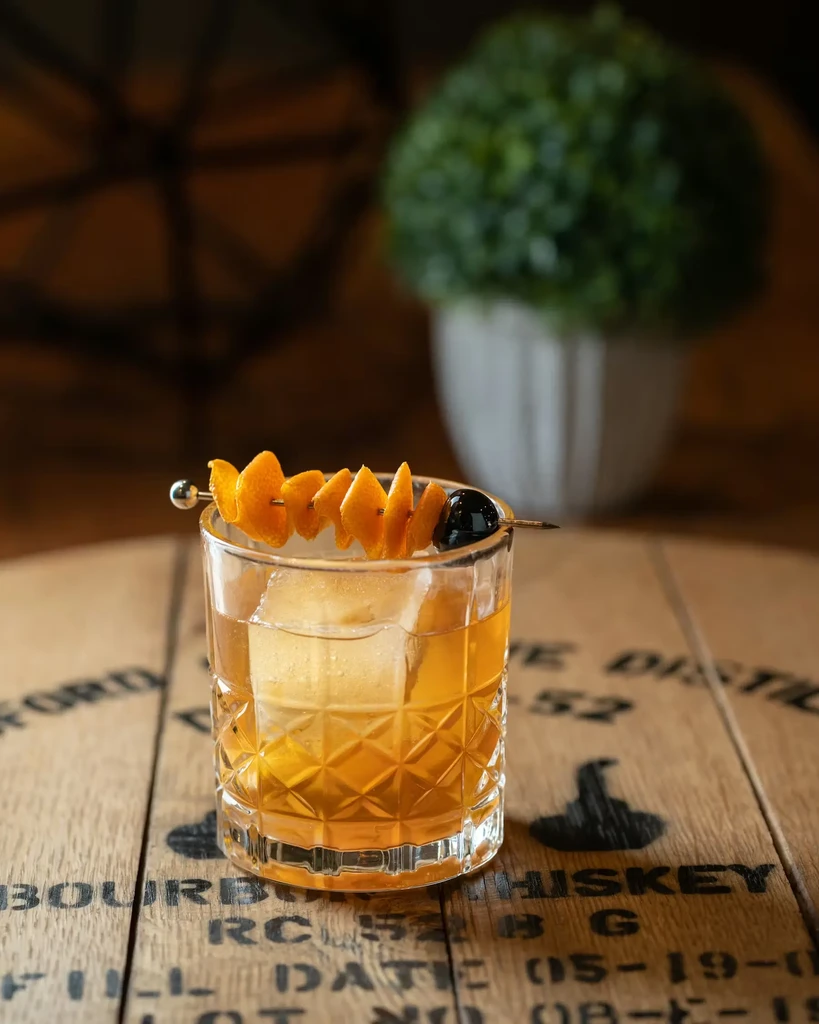 Amber whiskey cocktail with orange and cherry on a bourbon barrel.