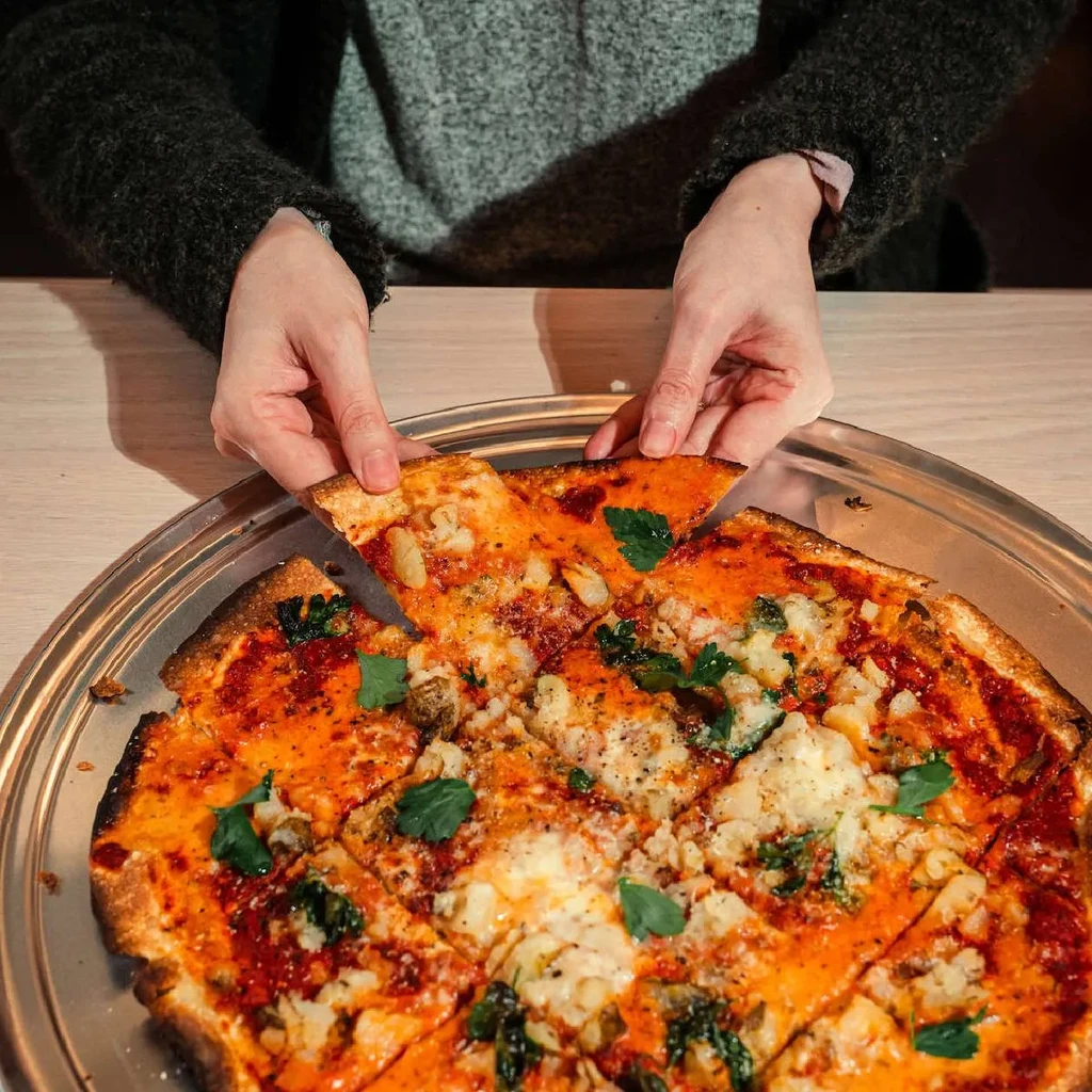 Hands lift a slice of sausage pizza with parsley from a metal tray.