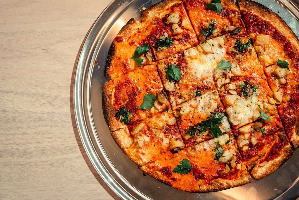 Square-cut pizza with potatoes, cheese, and herbs on a round metal tray.