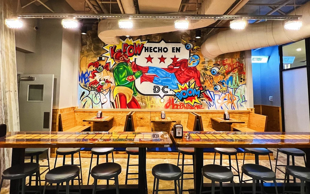 Colorful restaurant interior with a lucha libre mural, high-top bar, and booths.