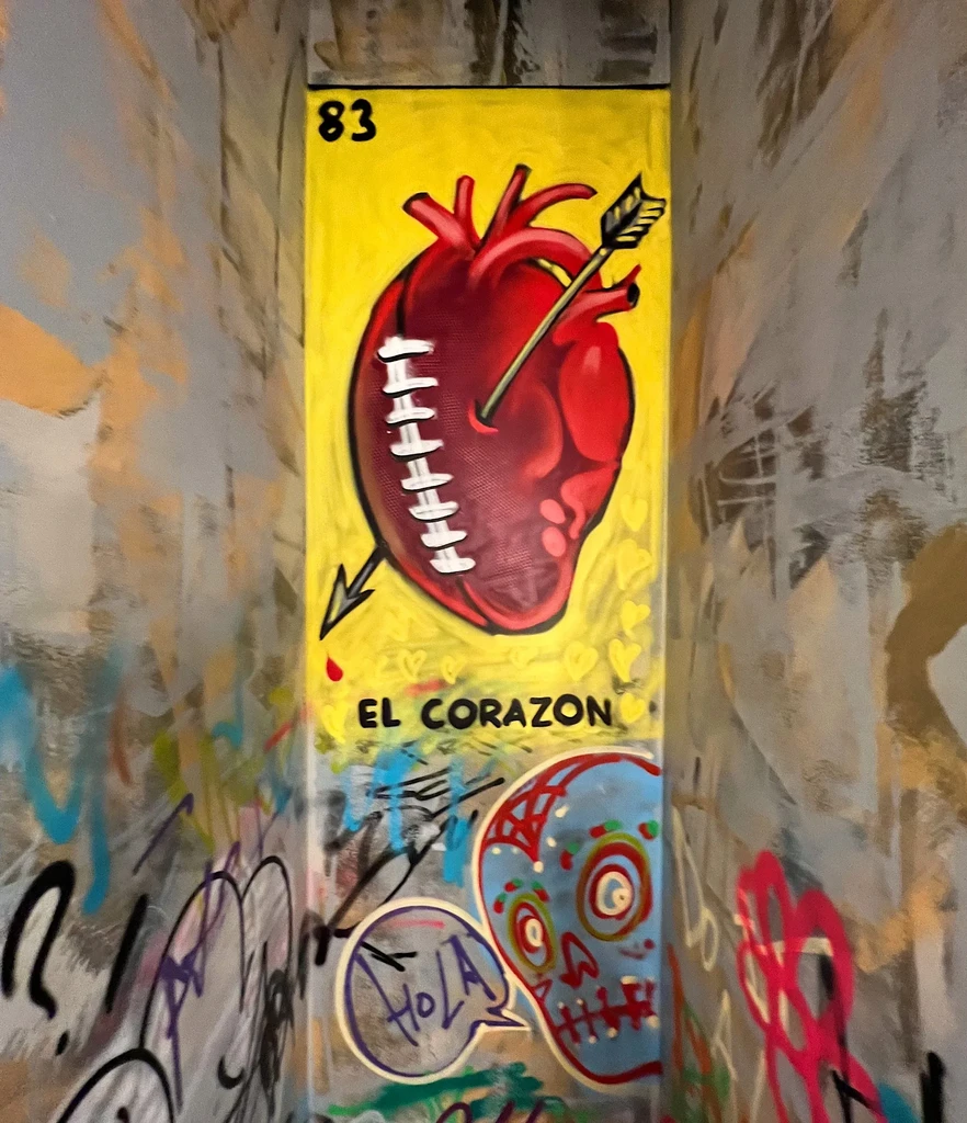 Vibrant interior wall with yellow Lotería-style heart art and colorful graffiti.