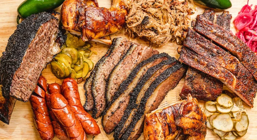 Overhead platter of barbecue brisket, chicken, pork, ribs, sausages, and pickles.