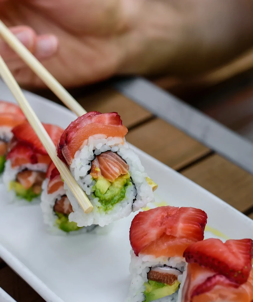Hand holding strawberry-topped sushi roll with salmon and avocado.