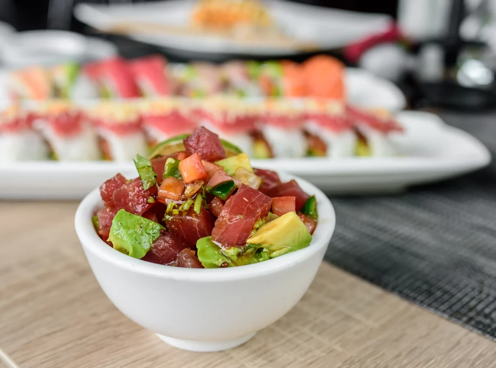 White bowl of tuna poke with avocado, sushi rolls on table.