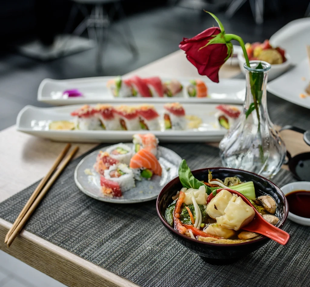 Close-up of sushi, dumpling soup, and a red rose on a table.