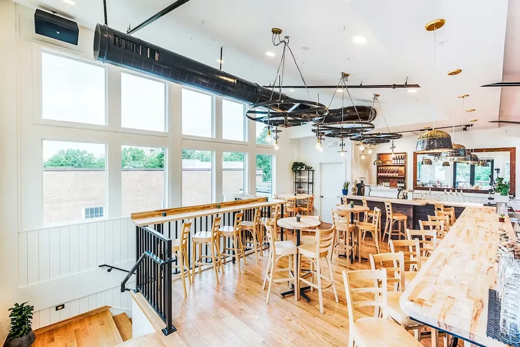 Modern rustic restaurant interior with exposed ductwork, wooden bar, tall windows.