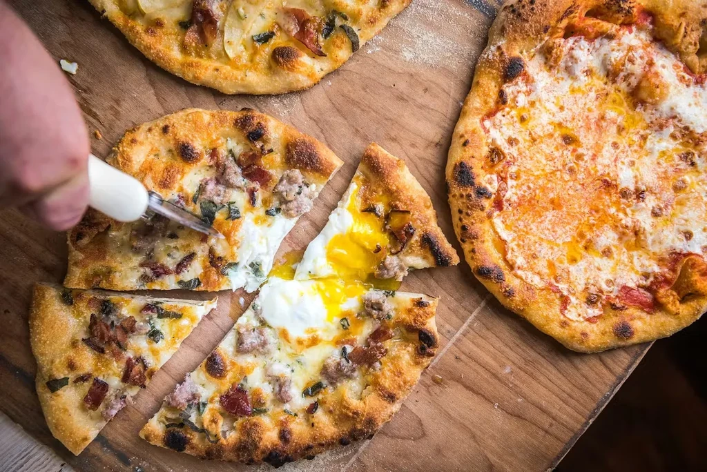 Hand slicing pizza with runny egg, next to two other pizzas.
