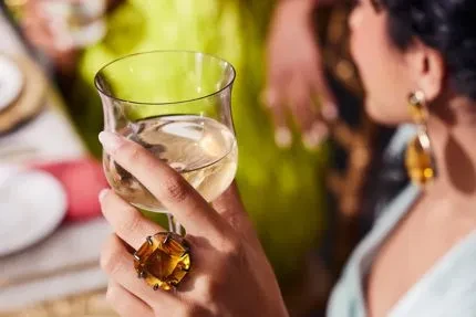 Hand with amber ring holds wine glass at social gathering.