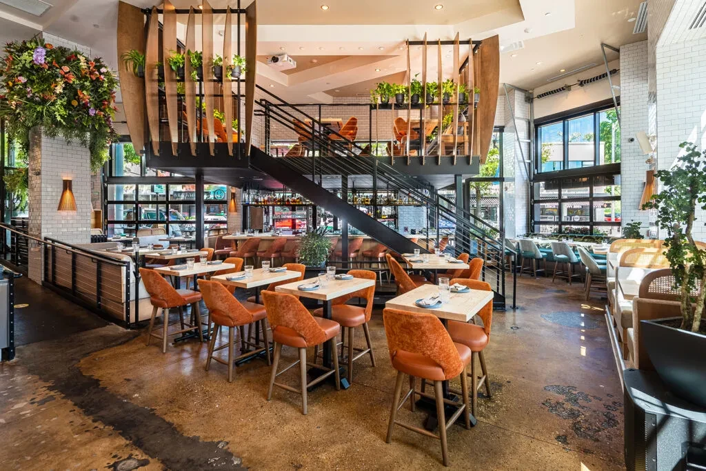 Modern multi-level restaurant with mezzanine, plants, concrete, and orange chairs.