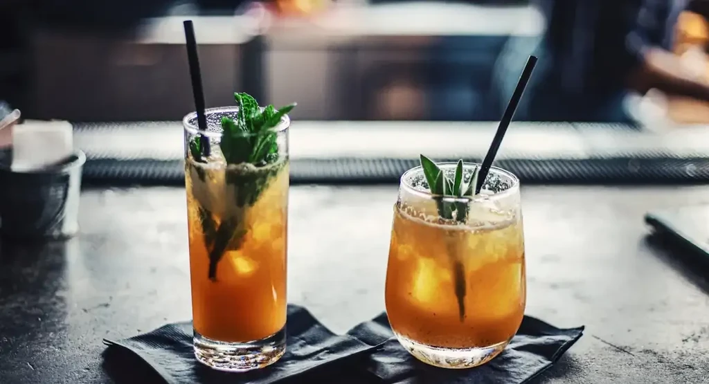 Two amber cocktails with ice and garnishes on a dark bar.