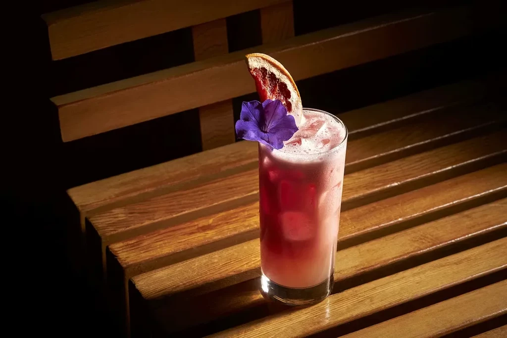 Vibrant pink cocktail with ice, dried orange, purple flower on wood.