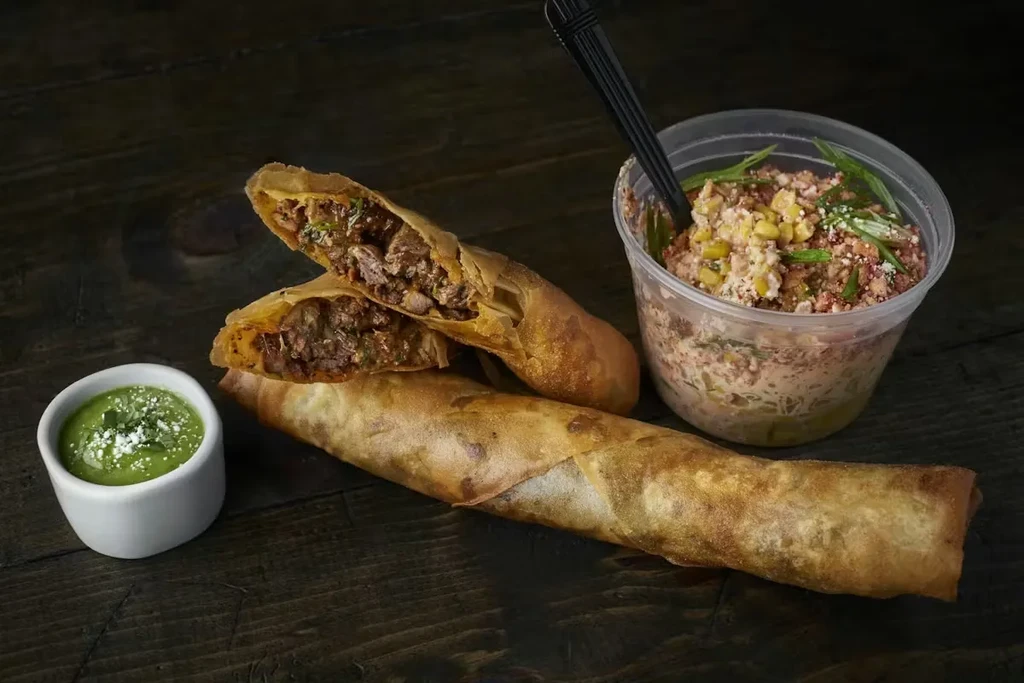 Fried taquitos, green sauce, and elote salad on a dark wooden surface.