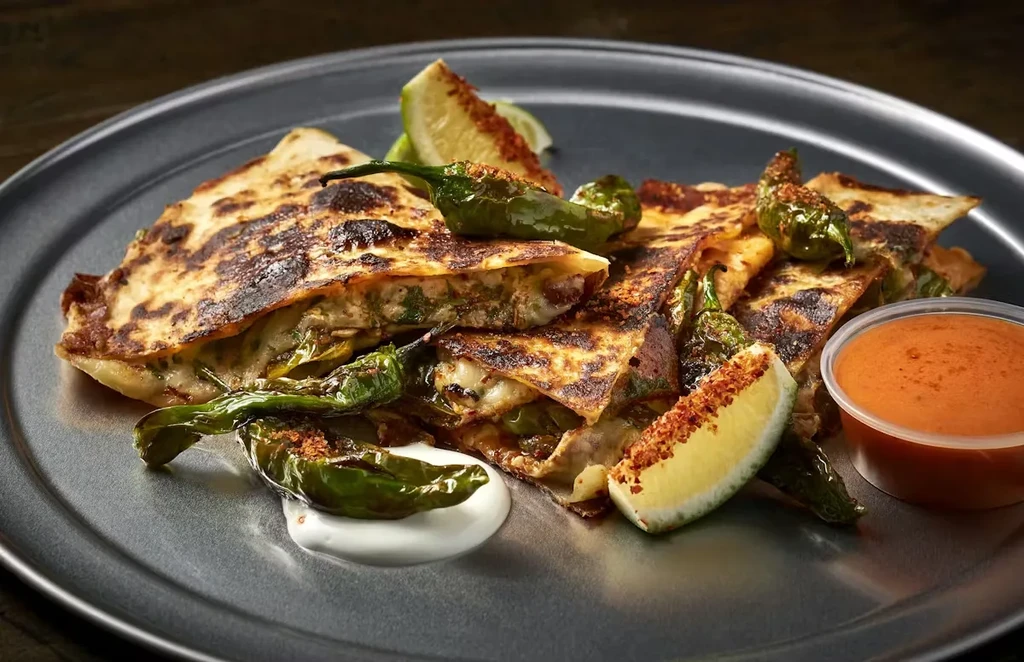 Grilled quesadilla triangles, charred peppers, lime, and sauces on a dark plate.