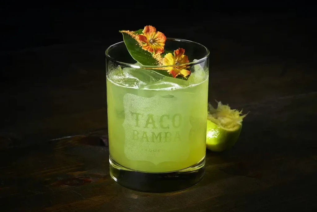 Vibrant green cocktail, floral garnish, ice, and "TACO BAMBA" glass.