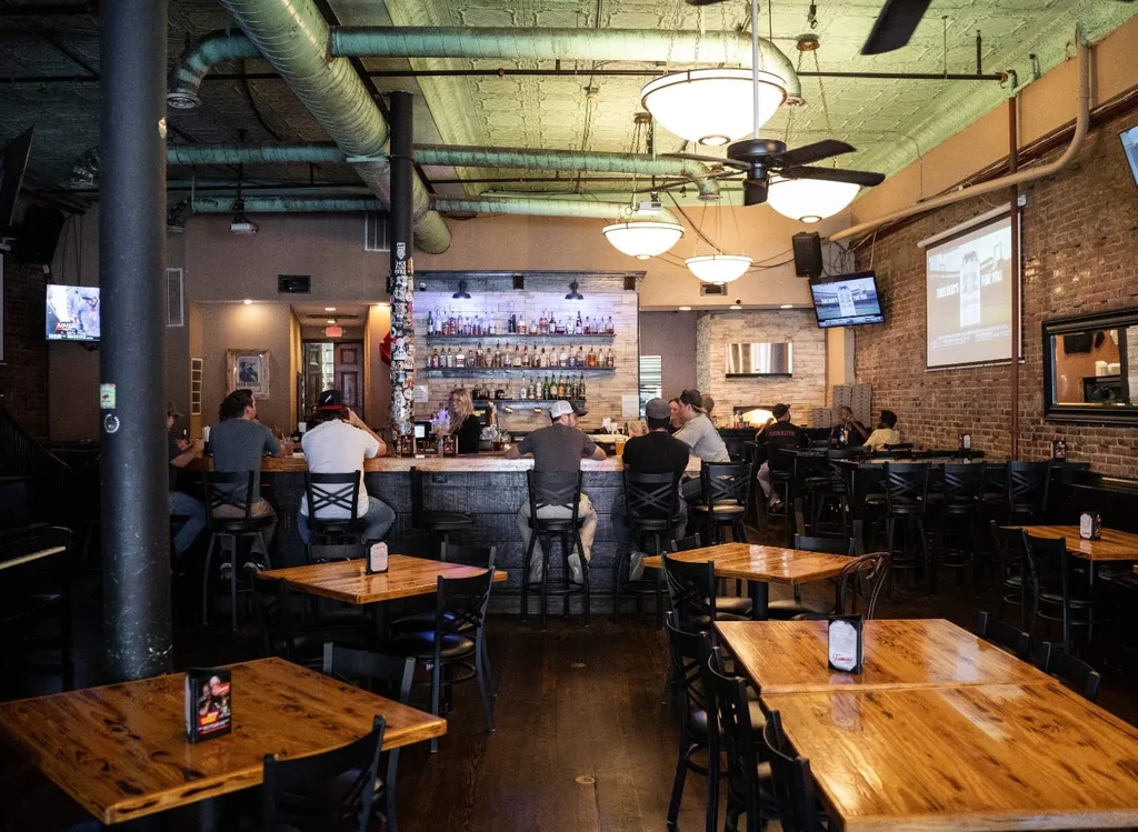 Industrial-chic restaurant bar interior with customers, exposed ducts, and brick walls.