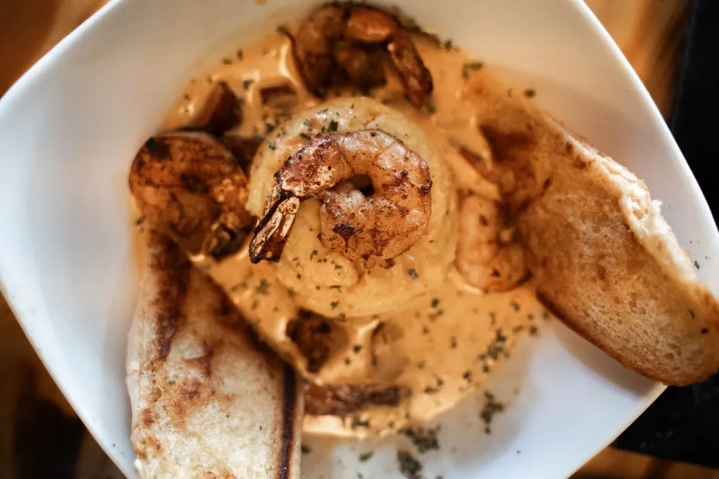 White bowl of shrimp and creamy grits with herbed sauce and bread.