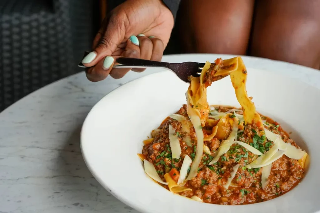 Fork lifting wide pasta with meat sauce, cheese, and herbs.