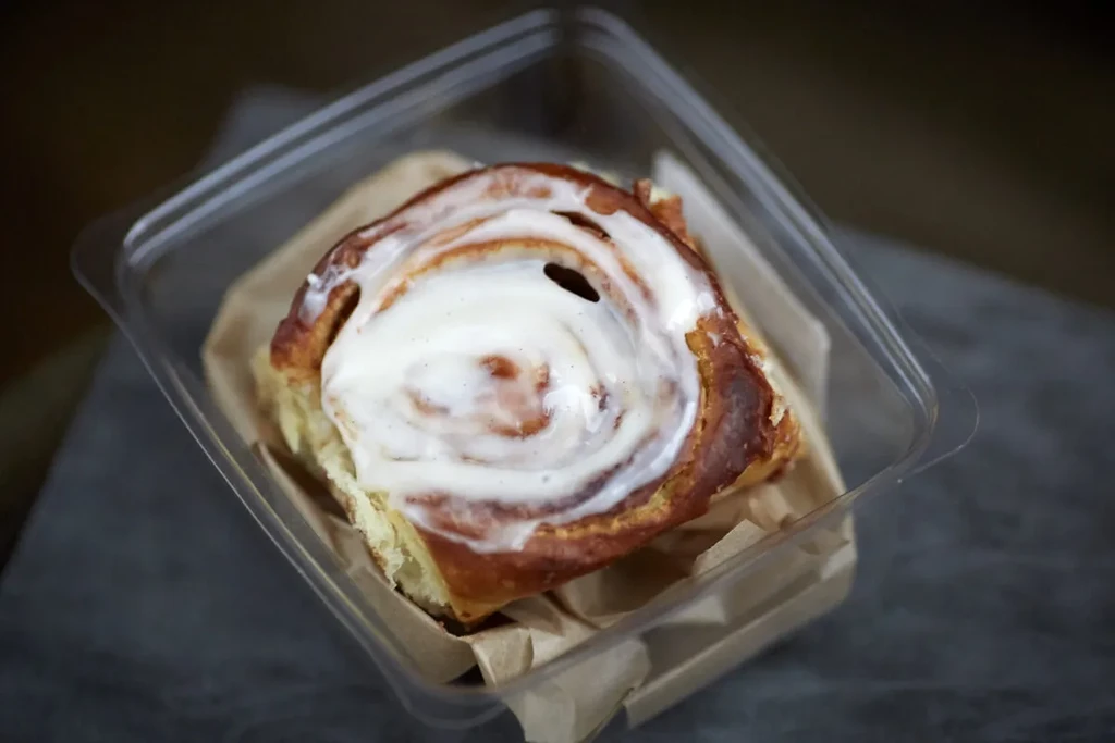 A single golden brown cinnamon roll with white icing in a clear container.