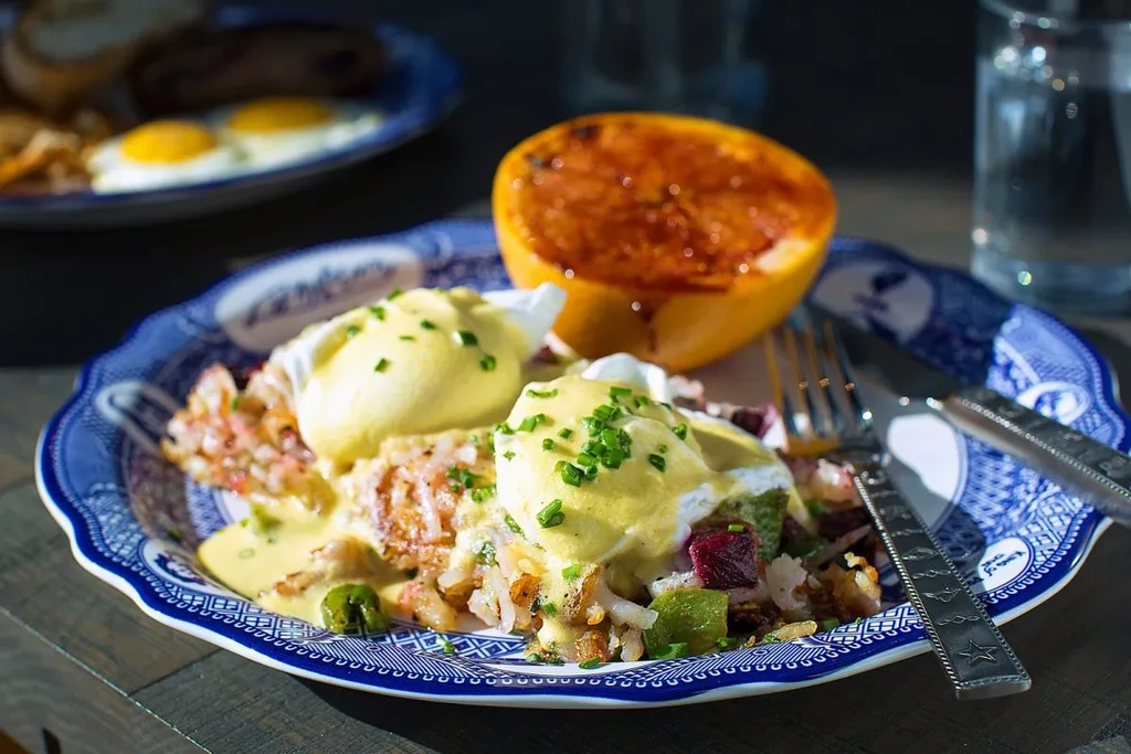 Eggs Benedict, hash browns, broiled grapefruit, and cutlery on patterned plate.