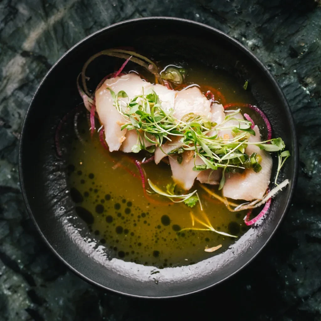 Sliced protein with microgreens, red garnish, and broth in black bowl.