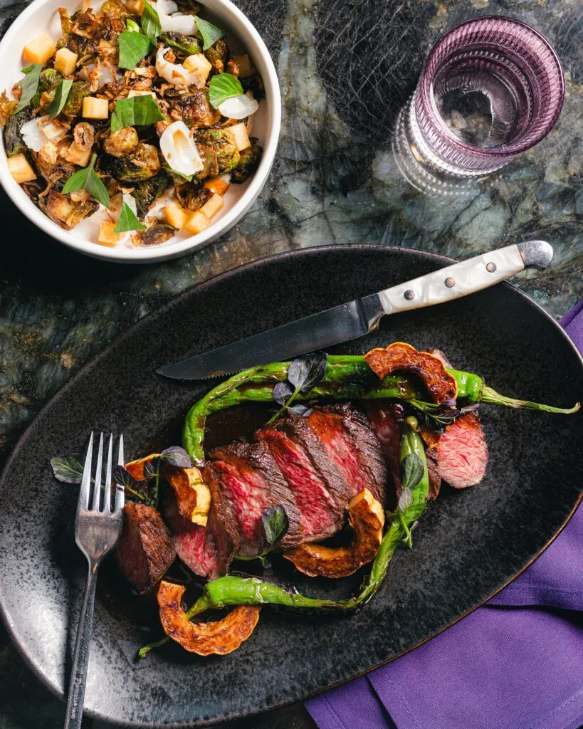 Overhead shot of medium-rare steak, vegetables, knife, fork on a dark table.