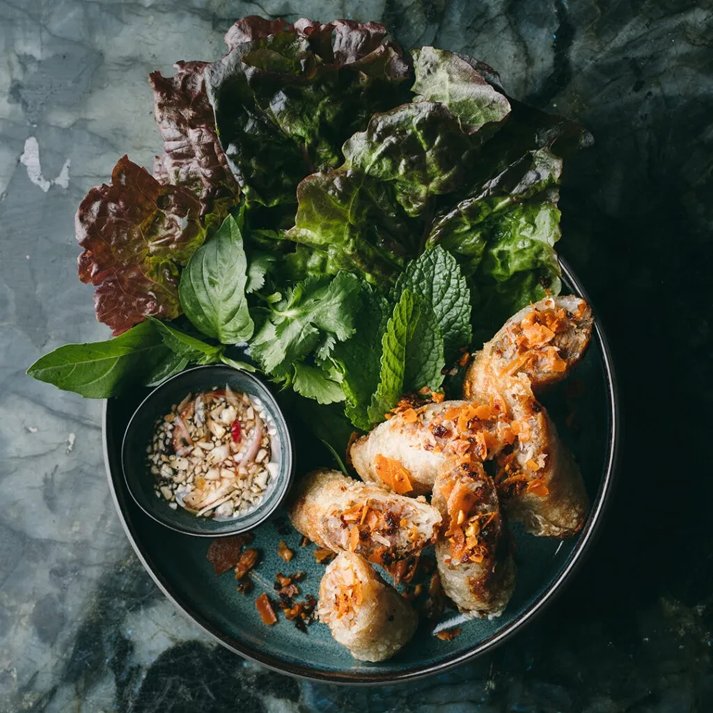 Golden fried spring rolls, fresh herbs, and dipping sauce on dark plate.