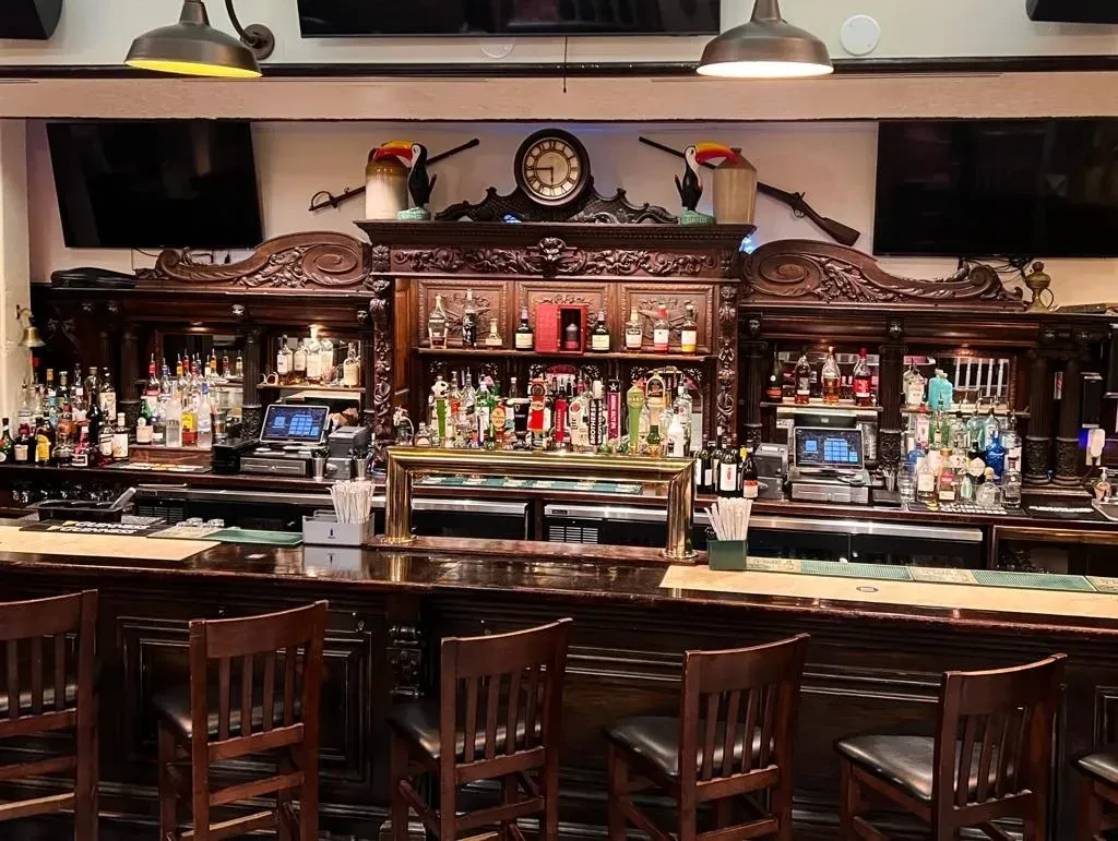 Elaborate dark wood bar with stocked backbar, clock, muskets, and TVs.