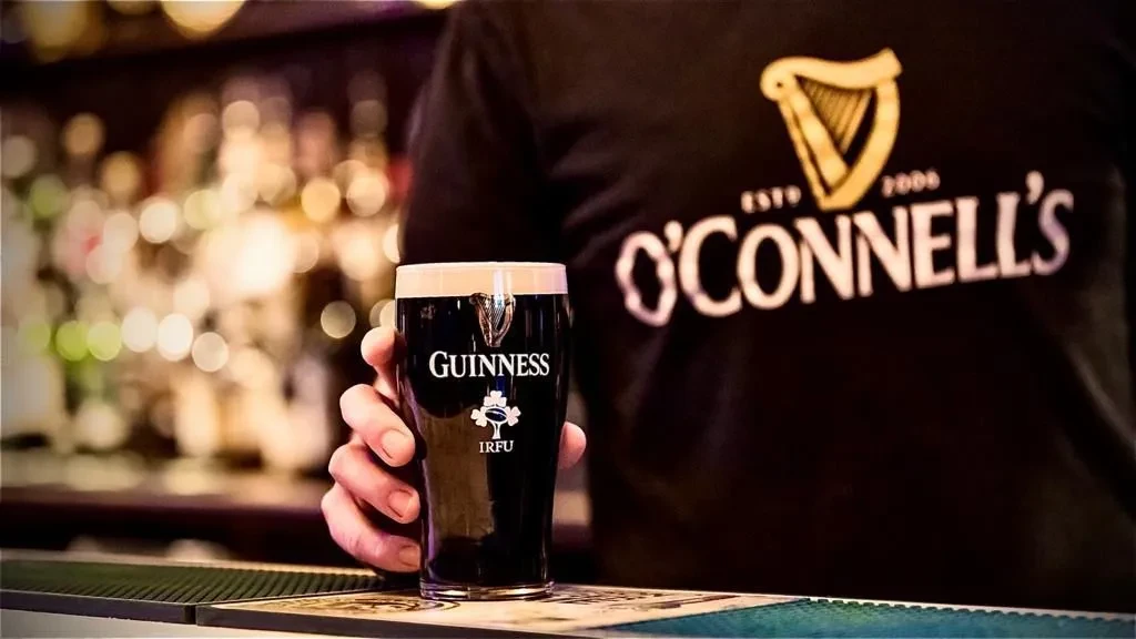 Hand in O'CONNELL'S shirt holds Guinness pint, IRFU logo, pub background.