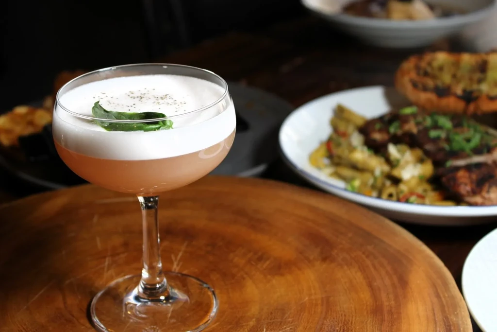 Two-toned cocktail with foam and green leaf on wood, blurred food background.