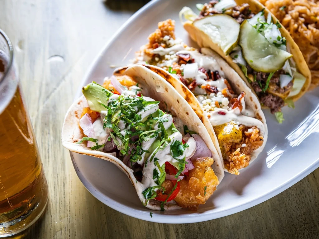 A plate of three distinct tacos and a glass of amber beer.