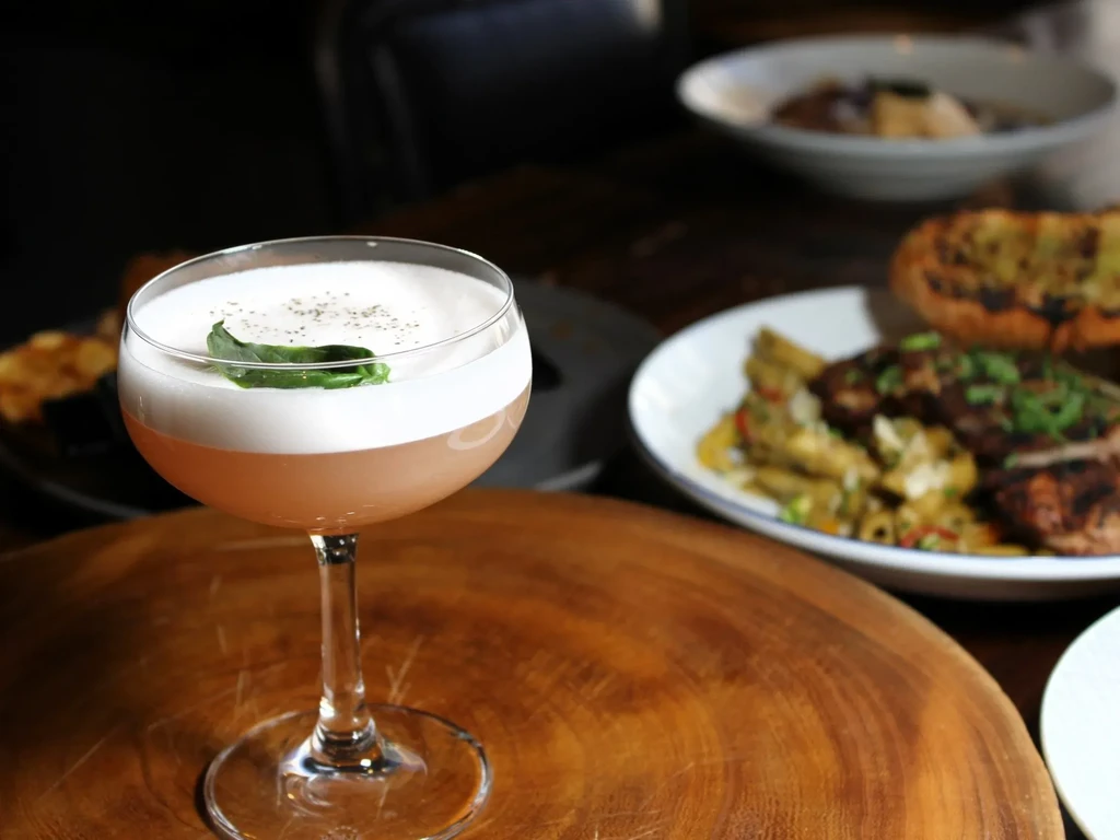Foamy cocktail with green leaf on a wooden table, blurred food.