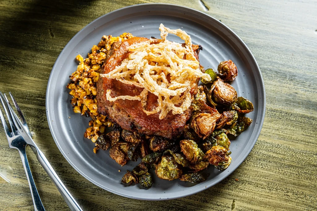 Plated roasted meat with crispy onions, corn, and Brussels sprouts.