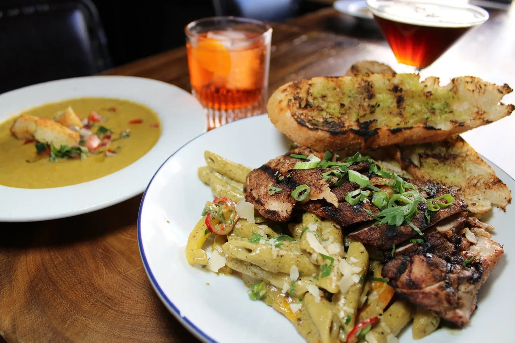 Top-down: creamy soup, pasta with grilled meat, garlic bread, two cocktails.
