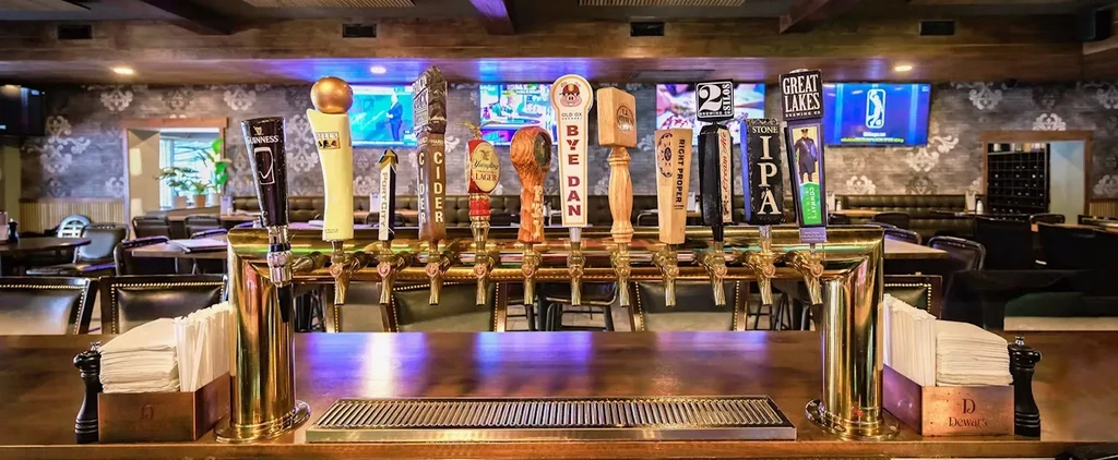 Many brass beer taps with brand logos on a wooden bar.