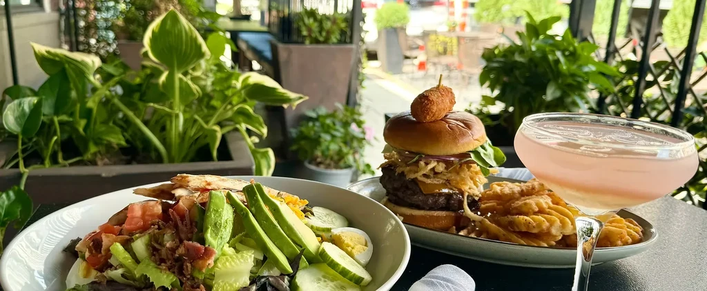 A colorful salad, large burger, and pink cocktail on an outdoor table.
