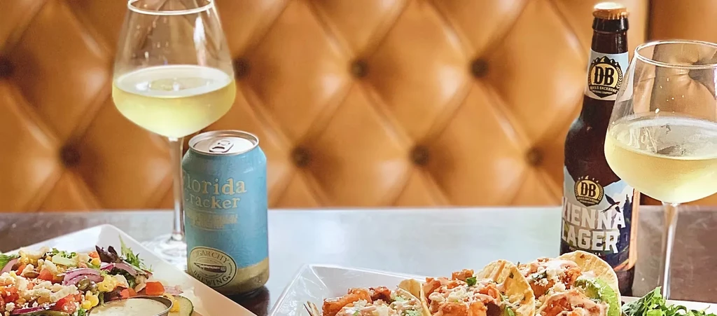 Tabletop scene with salad, tacos, beers, and wine against a tufted background.