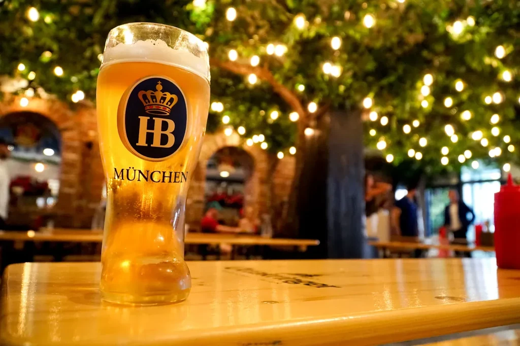 Golden HB München beer on a wooden table with string lights.