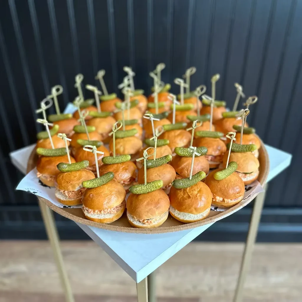 Tray of numerous sliders with light filling, pickles, and skewers.